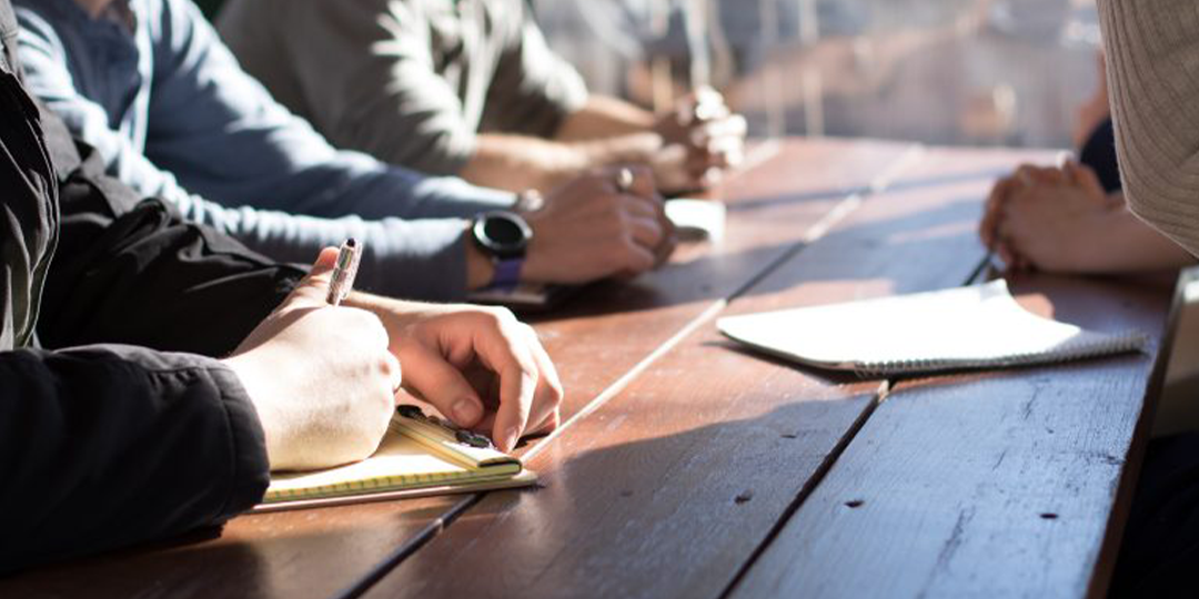 Image of people sitting around a table discussing while writing on paper and notebooks